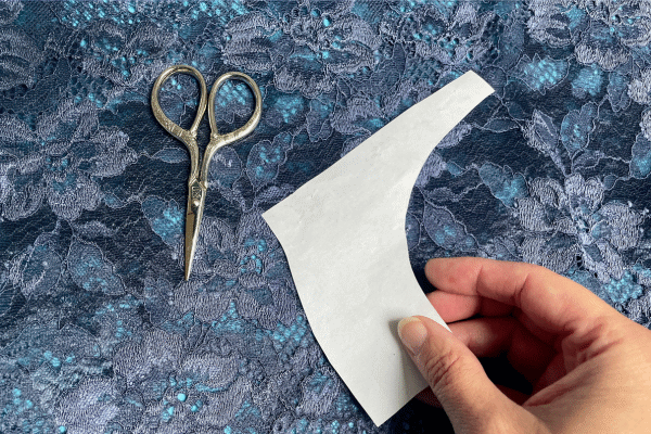 Eine Hand hält ein weißes Papierschnittmusterstück auf dunkelblauer, blumengemusterter Spitze. Daneben liegt eine kleine, filigrane Stoffschere. so sitzt deine Wäsche perfekt